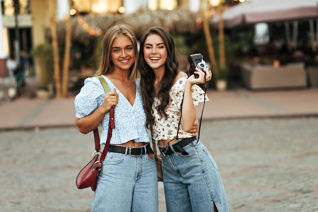 Tanned curly brunette woman in loose denim pants and floral blouse holds retro camera and poses with young girlfriend outside Pretty blonde girl in blue top and jeans smiles outdoors