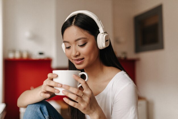 Tanned dark-haired woman drinks tea and listens to music with headphones while sitting in kitchen