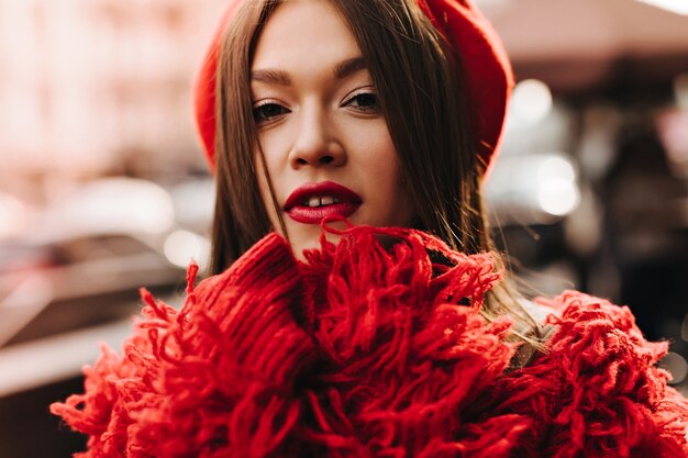 Tanned dark-haired woman in red wool coat and beret looking at camera against background of city street.