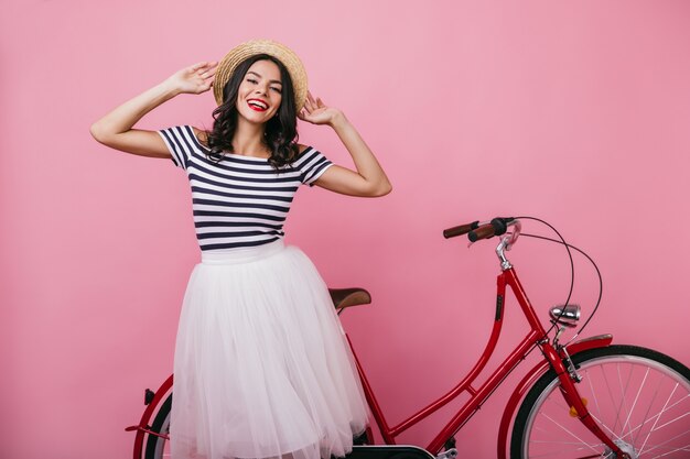 Tanned ecstatic woman touching her straw hat. relaxed caucasian girl posing with bicycle.