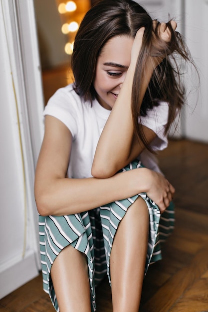 Загорелая девушка в милой пижаме смеется и играет со своими волосами Фото в помещении утонченной юной леди, сидящей на полу с искренней улыбкой