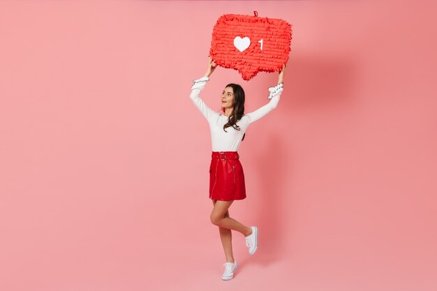 ファッショナブルな革のスカートの日焼けした女の子は、Instragramのような形の装飾でピンクの背景に跳ね返ります。