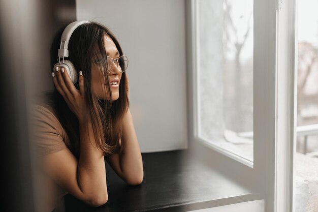 Tanned girl in light brown Tshirt and glasses leaned on windowsill listening to music with headphones and looking out window