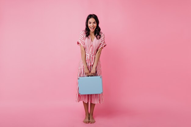 Tanned girl looks in surprise, modestly showing off her bright briefcase. Picture of European model in summer dress.