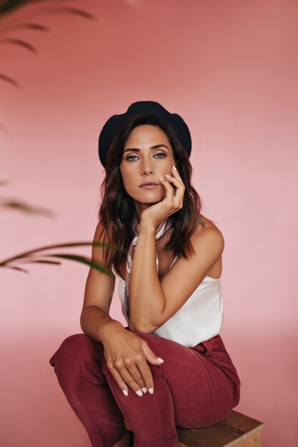 Tanned lady in beret posing on pink background. Brunette adult woman with wavy short hair in beret sits thoughtfully