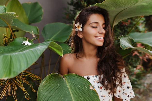 Tanned long-haired woman in white top with floral print enjoying summer day on vacation in warm country