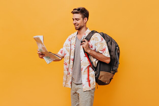 Загорелый турист в модном легком крутом наряде и солнечных очках позирует с рюкзаком и картой на изолированной оранжевой стене