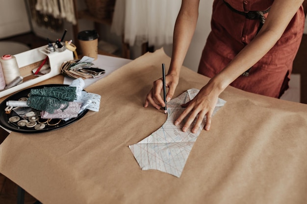 リネンの赤いドレスを着た日焼けした女性がクラフト紙に布のパターンを描くファッションデザイナーがオフィスで衣服のサンプルを作成する