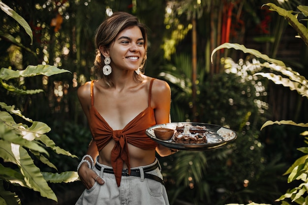 Tanned woman in shorts and brown bra smiles and holds board with waffle and nuts