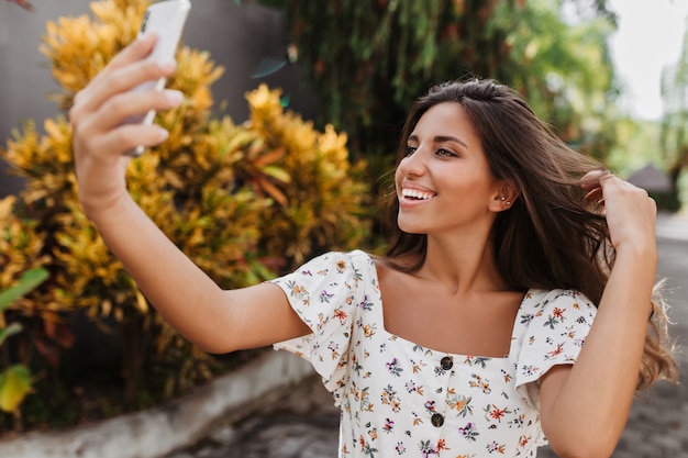 Tanned woman touches her long dark hair and takes selfie against wall of trees