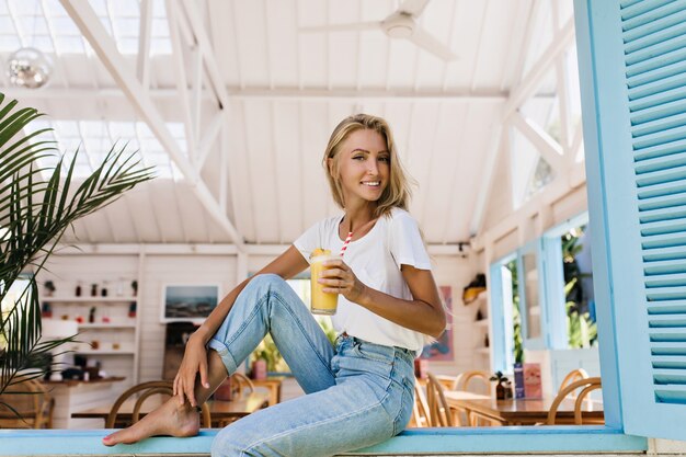 tanned woman in white t-shirt having fun in morning and drinking juice. Portrait of romantic blonde girl in casual clothes holding glass of fruit cocktail and smiling.