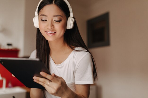 Tanned woman in white T-shirt listens to songs in headphones and looks at screen of tablet