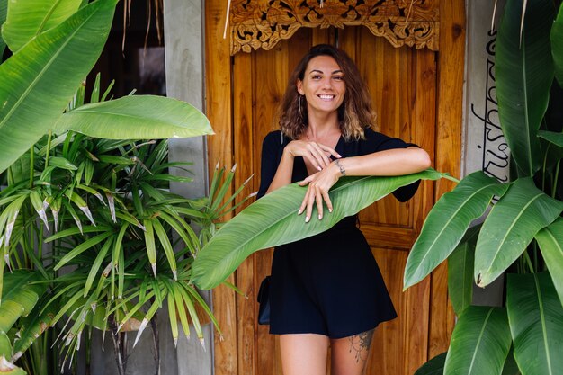 Tanned woman with short curly hair in black sexy overalls by door of tropical villa on vacation at sunset light