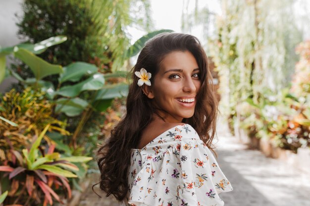 Tanned woman with white flower in wavy dark hair smiles while walking in tropical park