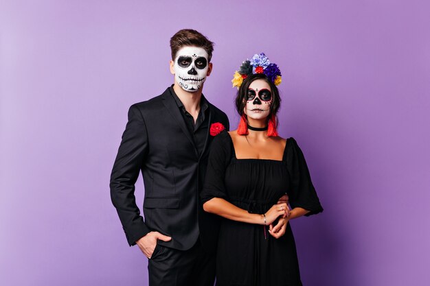 Tanned young woman in zombie costume enjoying photoshoot with boyfriend. Funny couple in halloween attires posing 