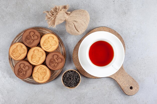 Tart cookies and a cup of tea on wooden boards next to a small bowl of tea leaves and a sack on marble background. High quality photo