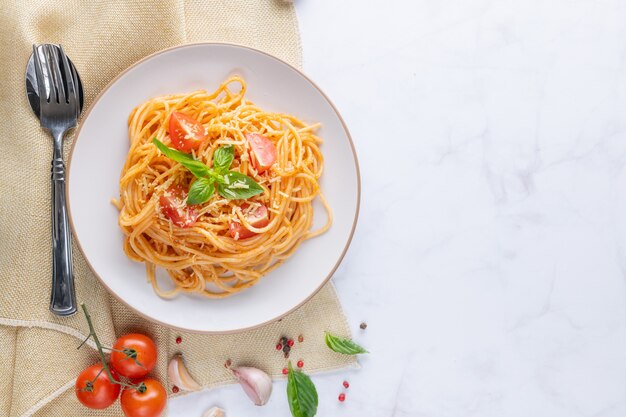 Tasty appetizing classic italian spaghetti pasta with tomato sauce, cheese parmesan and basil on plate and ingredients for cooking pasta on white marble table. Flat lay top view copy space.