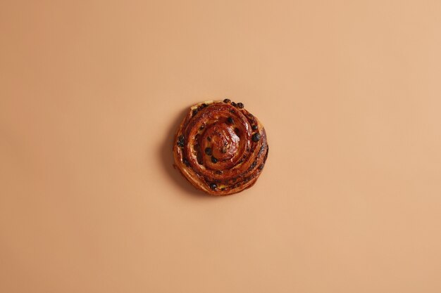 Tasty appetizing puff round spiral soft bun with raisins baked in bakery. High calories bakery product containing much fat and sugar. Homemade roll on beige studio background. Sweet food concept.