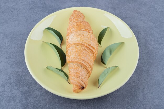 Tasty buttery croissant with leaves on yellow plate.