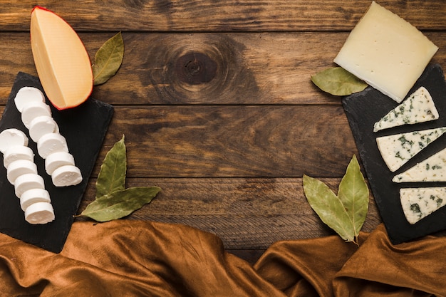 Tasty cheese on black slate rock with bay leaves and silk textile over wooden surface