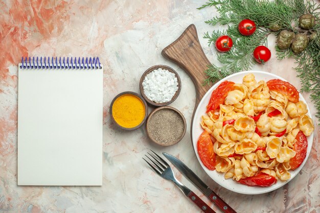 Tasty conchiglie on a white plate on wooden cutting board and knife different spices and notebook on mixed color background