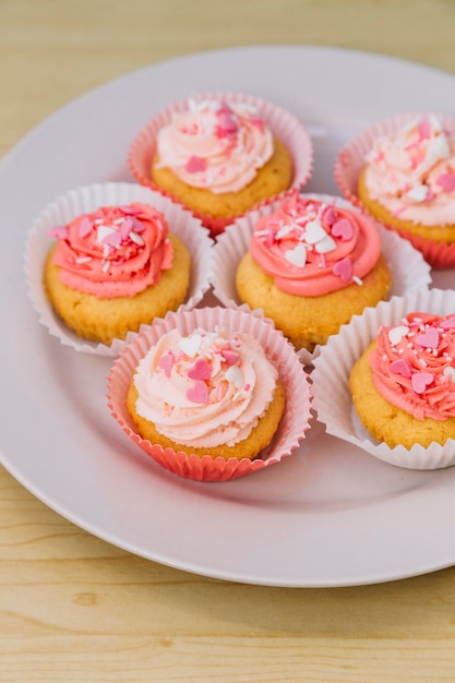 Tasty cupcakes with cream and sprinkles on white plate over the wooden desk