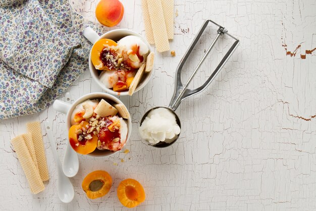 Вкусное свежее холодное аппетитное ванильное мороженое с орехами, абрикосами, вафлями и сиропом на белом столе с ингредиентами для приготовления десерта. Вид сверху.