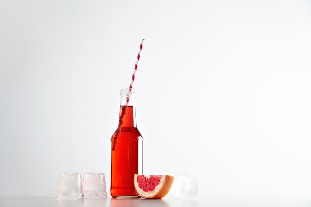 Tasty fresh grapefruit lemonade in transparent bottle with striped red drinking straw near ice cubes and pomelo slice isolated on white