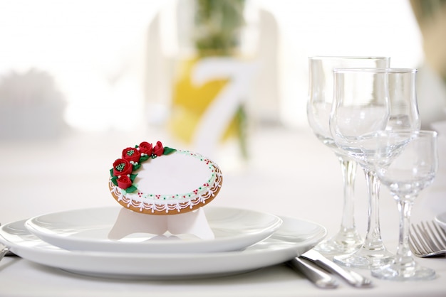 Tasty gingerbread cookie covered with glaze and decorated with tiny red roses and pattern stands on the festive wedding table with glasses and other dishes. Looks delicious and cute.