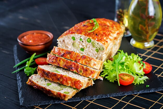 Tasty homemade ground baked chicken meatloaf with green peas and broccoli on black table.