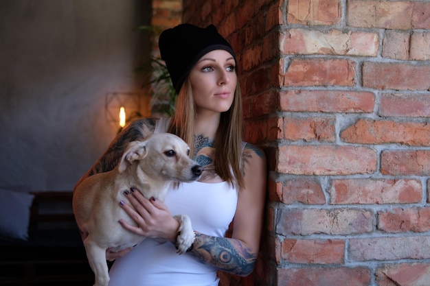 無料写真 tattoedブロンドの女性は、目をそらして、レンガの壁に寄りかかっている間、彼女の犬を保持します。
