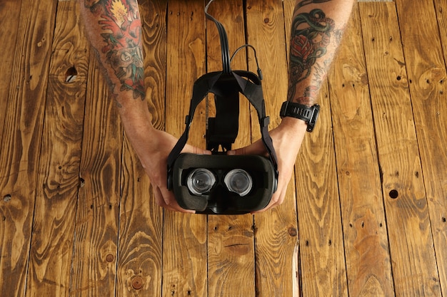Tattoed hands hold vr glasses upside down, presentation of new technology, isolated on rustic wooden board