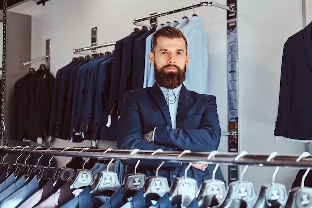 Татуированный мужчина со стильной бородой и волосами, одетый в элегантный костюм, стоит со скрещенными руками в магазине мужской одежды.