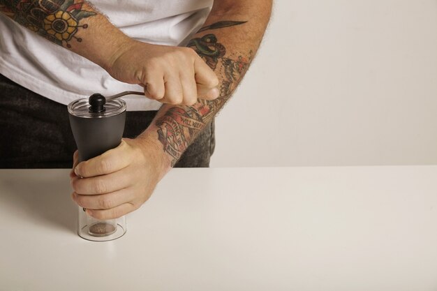 A tattoed man in white t-shirt and black jeans grinds coffee beans in a modern slim manual burr grinder, close up