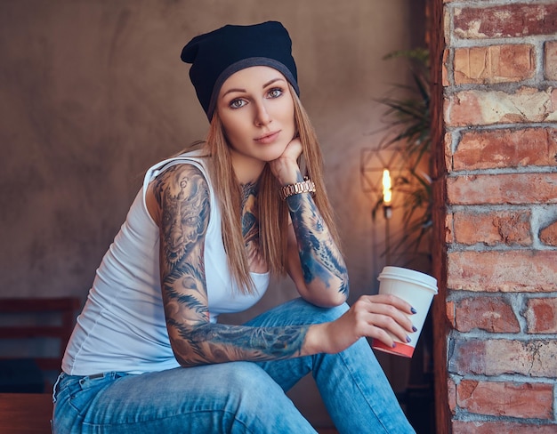 Foto gratuita una bionda sexy tatuata con una maglietta e un cappello con una tazza di caffè in una stanza con interni soppalcati.