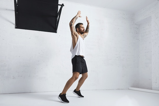 Бесплатное фото Татуированный и мускулистый спортсмен, выполняющий прыжки, изолирован на белой кирпичной стене рядом с черной перекладиной, глядя вправо