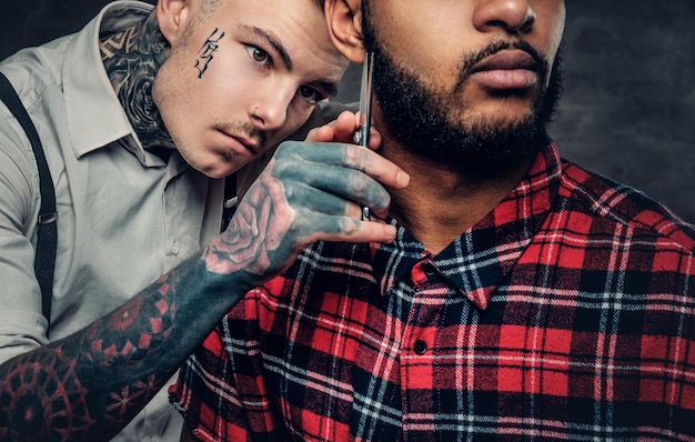 Татуированный парикмахер стрижет бороду чернобородому мужчине.