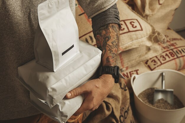 Tattooed barista holds blank package bags with freshly baked coffee beans ready for sale and delivery