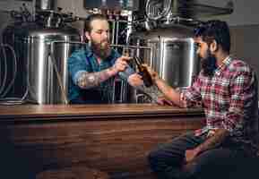 Free photo tattooed bearded caucasian and indian men presenting craft beer in the microbrewery.