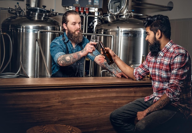 Татуированные бородатые кавказцы и индийцы представляют крафтовое пиво в пивоварне.