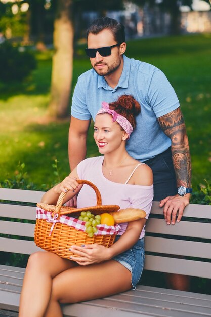 Tattooed bearded male and redhead female are having a picnic on a bench in a park.