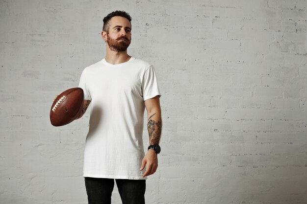 Tattooed and bearded model in plain white shortsleeve t-shirt holding a leather football on gray wall