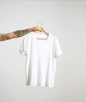 tattooed biker hand holds hang with blank white t-shirt from premium thin cotton, isolated on white