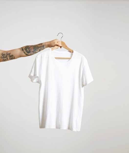 Tattooed biker hand holds hang with blank white t-shirt from premium thin cotton, isolated on white