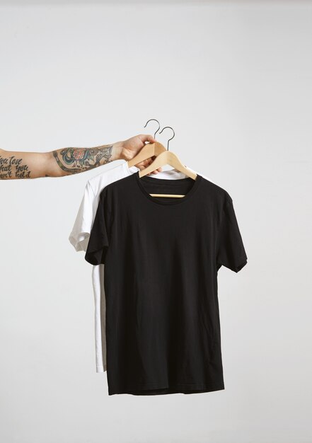 Tattooed biker hand holds wooden hangs with blank black and white t-shirts from premium thin cotton, isolated on white