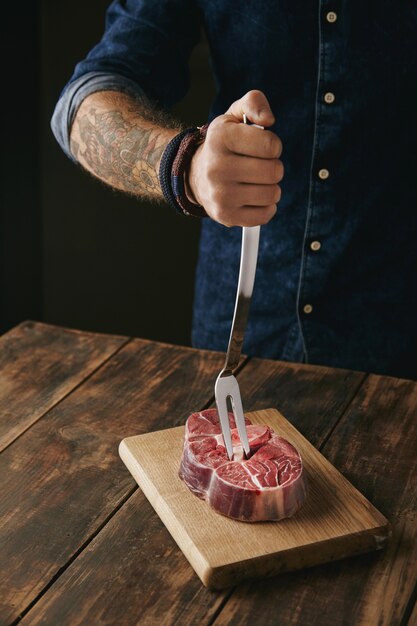 Tattooed butcher hand holds meat fork in fresh raw steak for dinner, unrecognizable