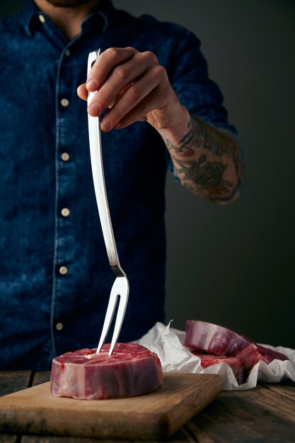 tattooed butcher hand holds meat fork in fresh steak for dinner, unrecognizable