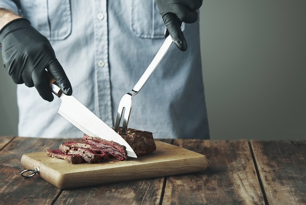 Tattooed butcher hands in black gloves with knife slice piece of grilled meat on wooden board