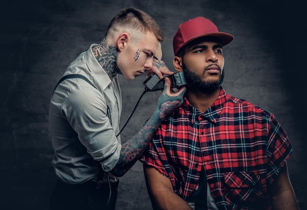 문신을 한 백인 남성 이발사가 세련된 흑인 남성에게 수염을 자르고 있습니다.