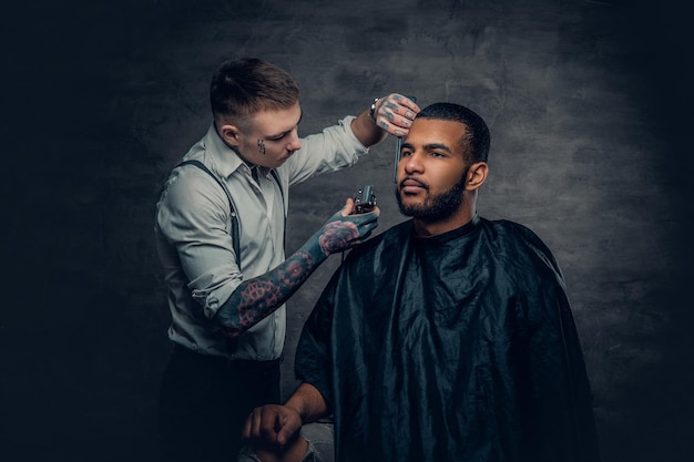 Tattooed Caucasian male barber cutting the beard to Black stylish man.